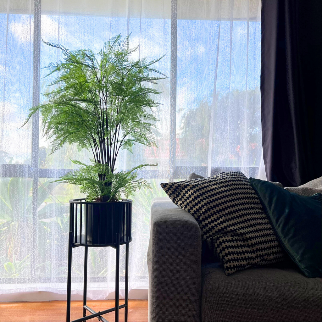 An artificial Rabbit's Foot Fern plant in a black pot, placed on a side table next to a gray sofa with cushions, with a large window in the background.