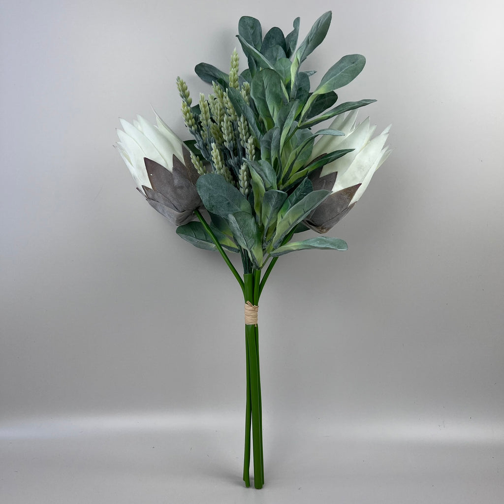 An artificial floral arrangement featuring white protea-like flowers, dusty blue-green leaves, and green stems, wrapped with a beige band at the base and photographed against a neutral gray background.