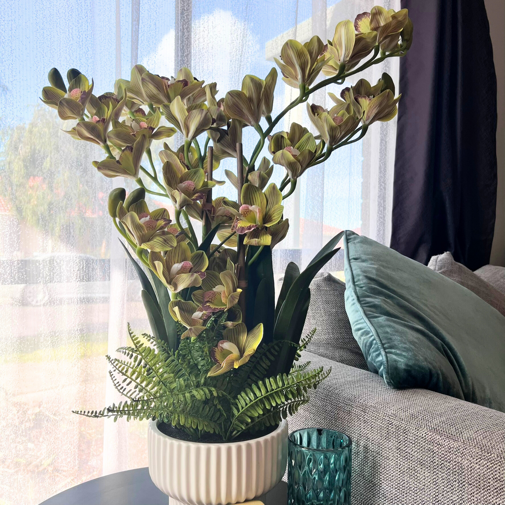 Yellow artificial cymbidium orchid in a white vase with ferns on a table 