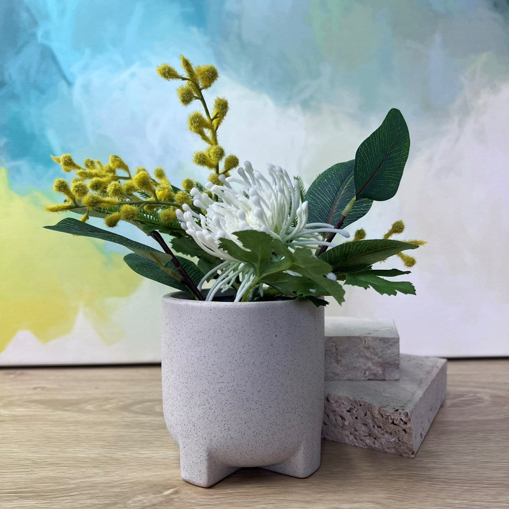 A small potted artificial floral arrangement in a speckled light gray ceramic pot, placed on a wooden surface with stacked stone blocks and a colorful abstract painting in the background.