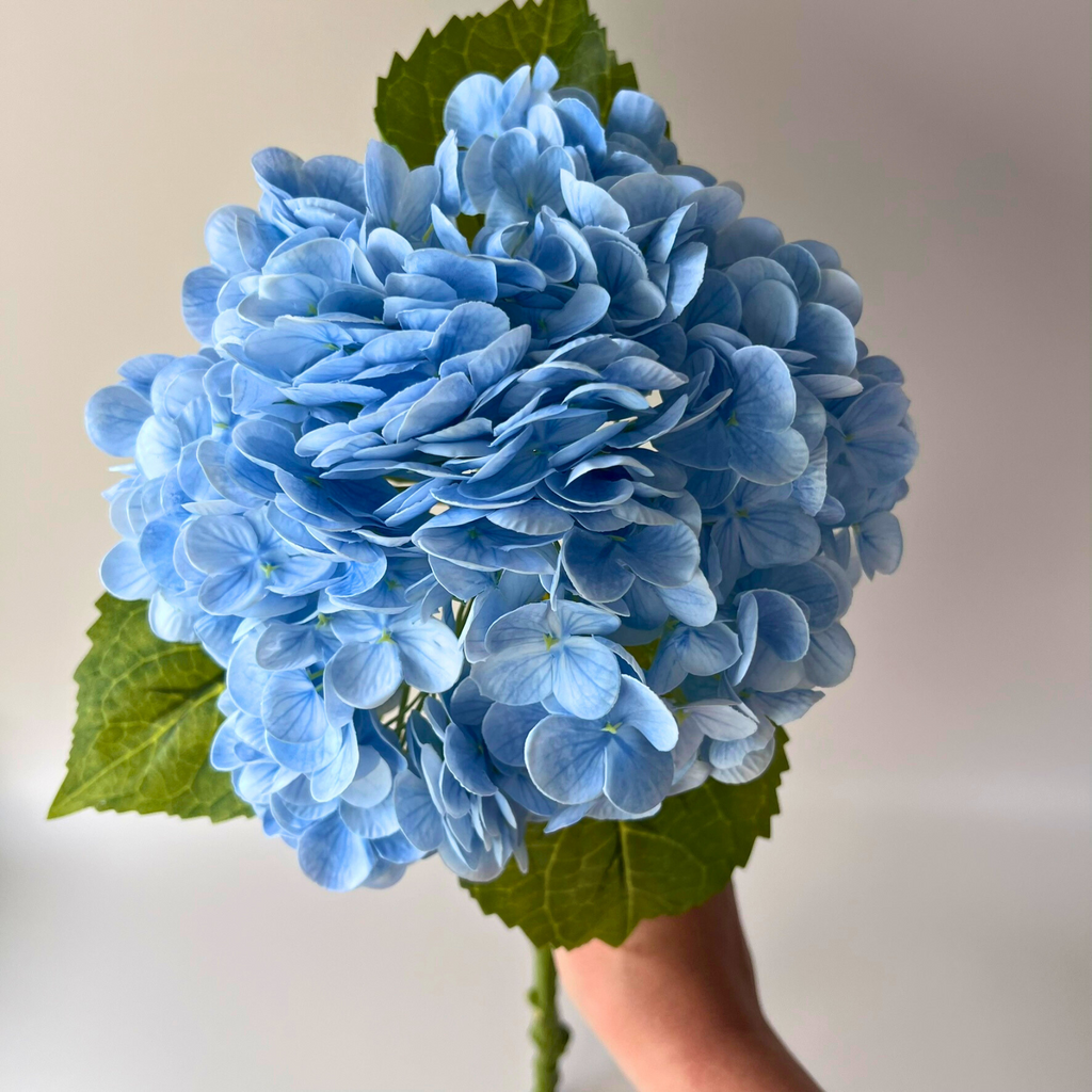 Artificial blue hydrangea held in a hand by the stem, showcasing beautiful blossoming blooms 