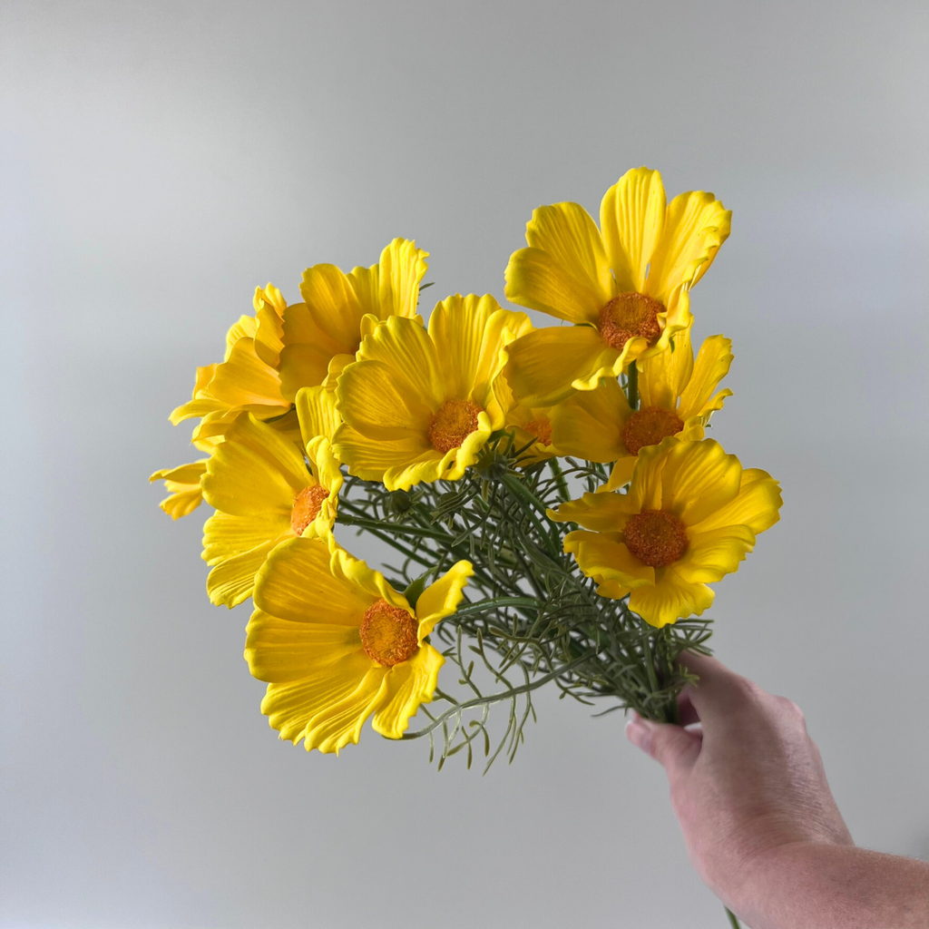 Hand holding a bouquet of faux yellow Cosmos blooms with green foliage at an angle against a plain background