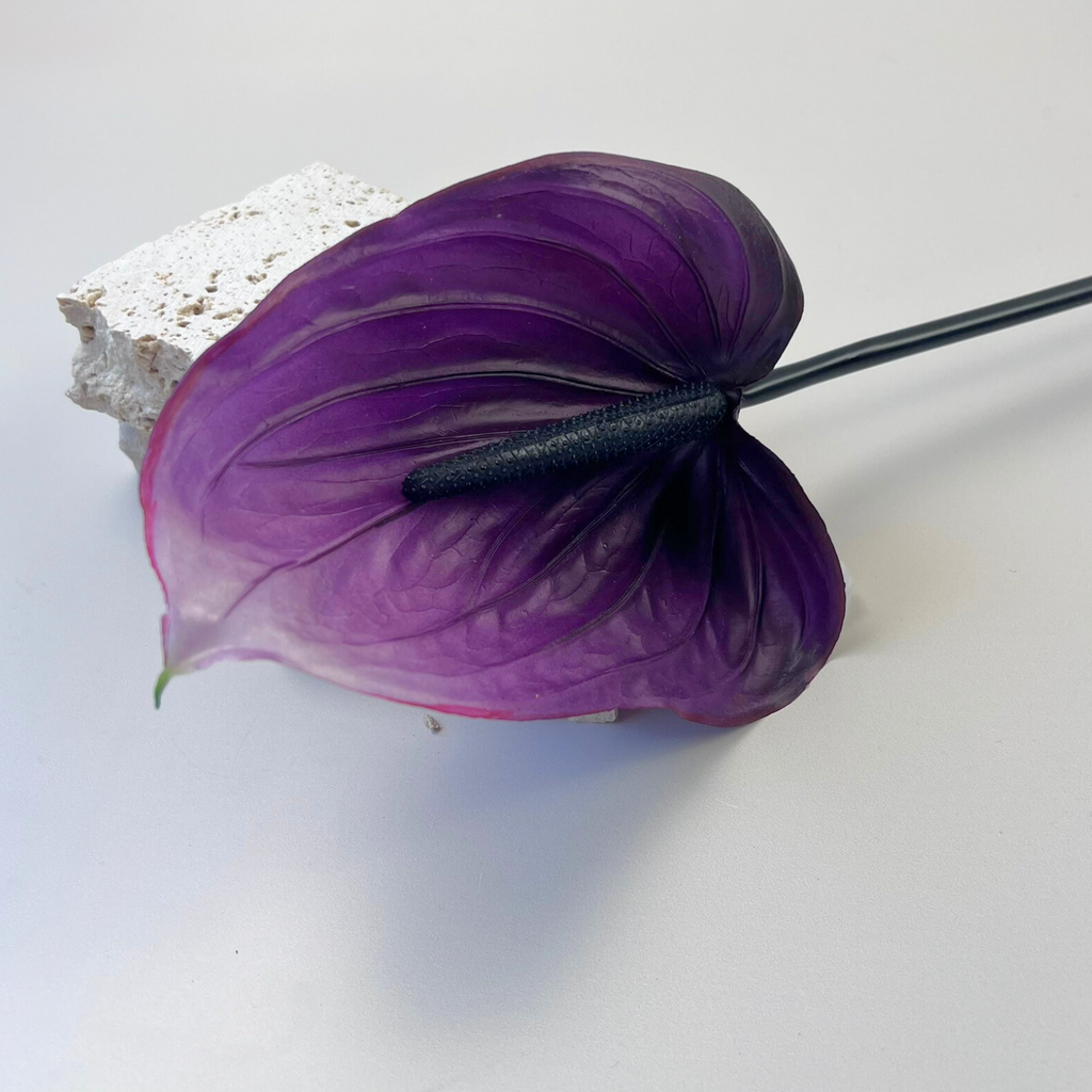 A dark purple artificial Anthurium laying next to decorative stones on a plain background