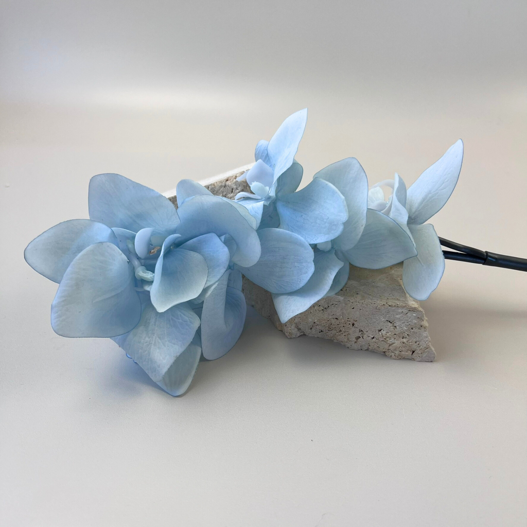 An artificial Phalaenopsis Orchid Stem with baby blue blooms styled on decorative stones against a plain background
