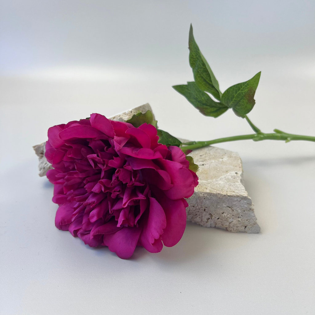 A Real Touch Peony stem with lifelike magenta petals and green foliage resting on decorative stones against a plain background